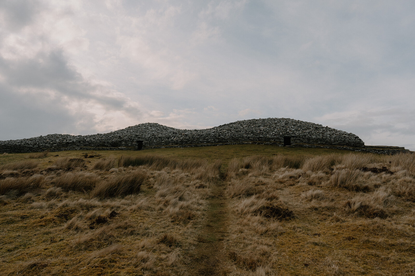A long chambered cairn of stacked stone rises from open moorland, a narrow path leading to its low entrance.