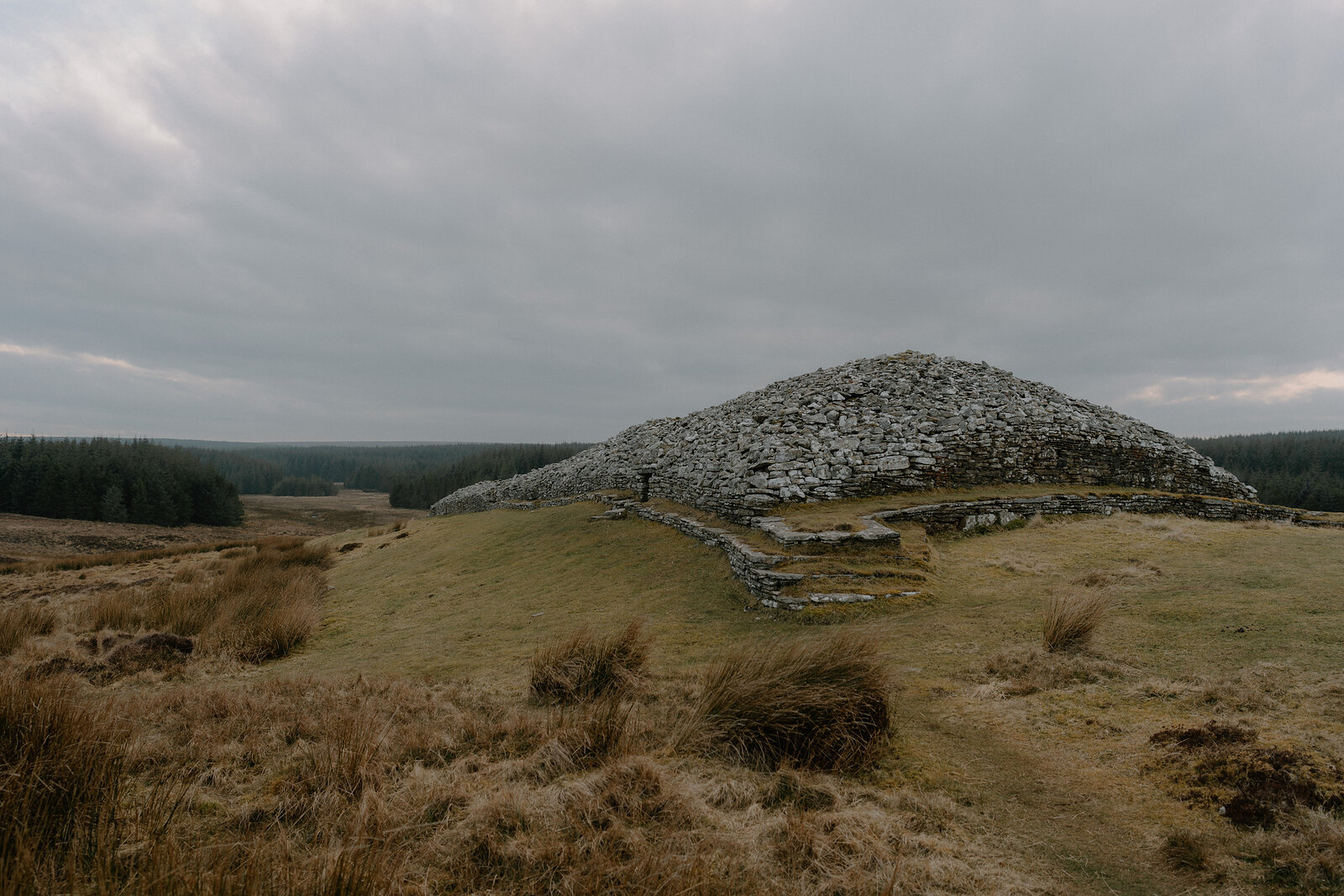 A large chambered cairn built of stacked stone rises from a grassy moor, a low entrance passage and dry-stone wall at its base.