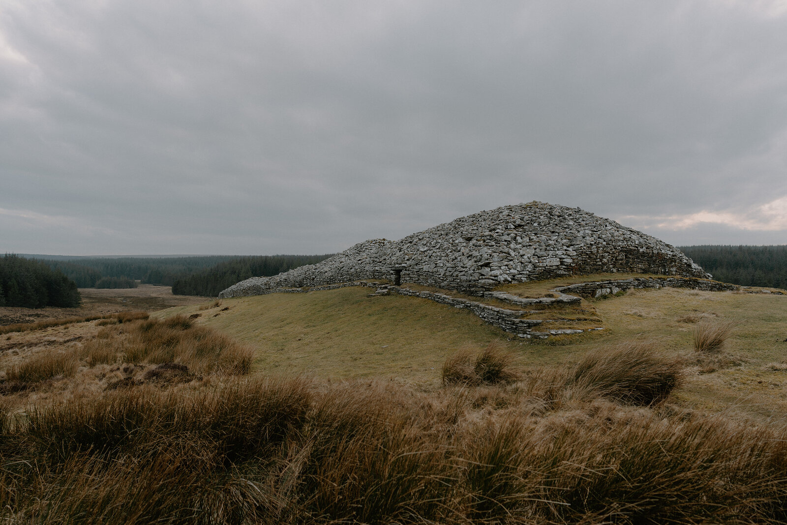 A large chambered cairn built of stacked stone rises from a grassy moor, a dark entrance passage set into its base.