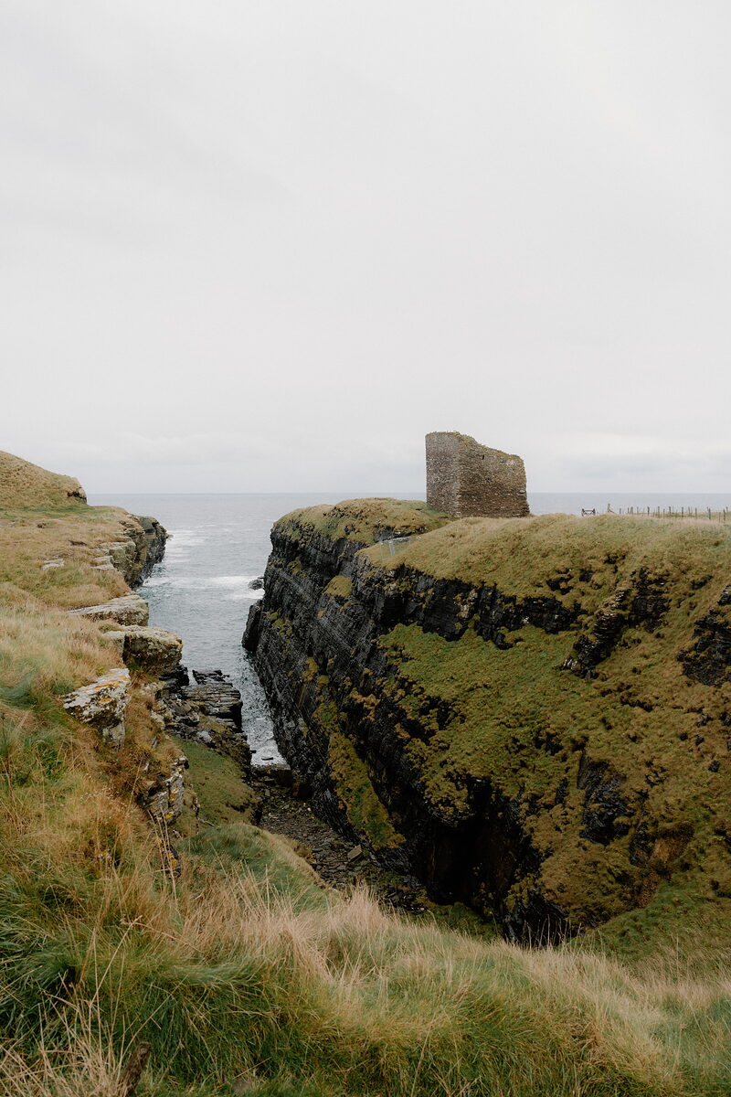 A ruined stone tower stands at the cliff edge above a narrow sea channel, waves breaking below grassy headlands on either side.