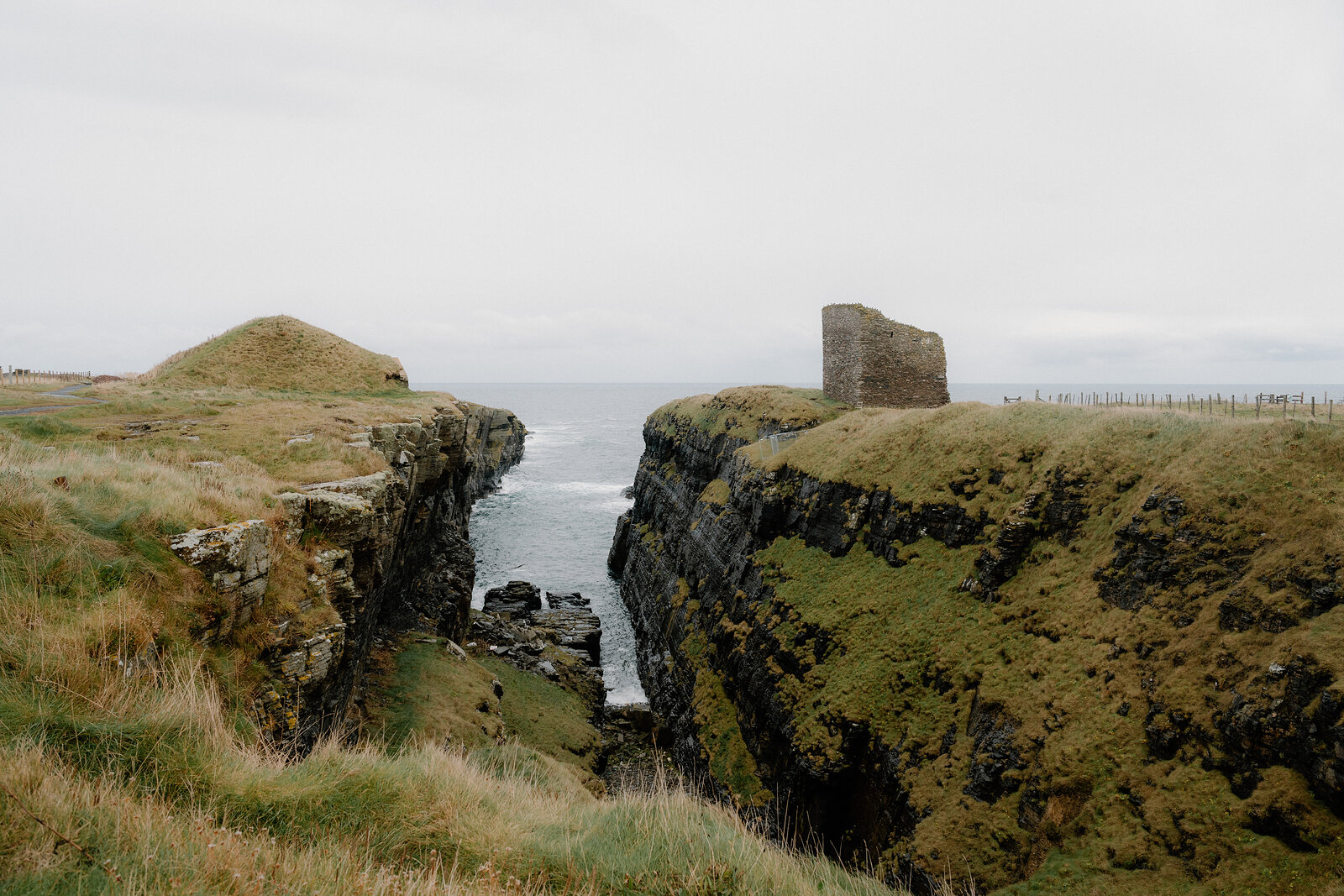 A ruined tower stands at the cliff edge above a narrow sea inlet, grassy headlands rising on either side of the water.