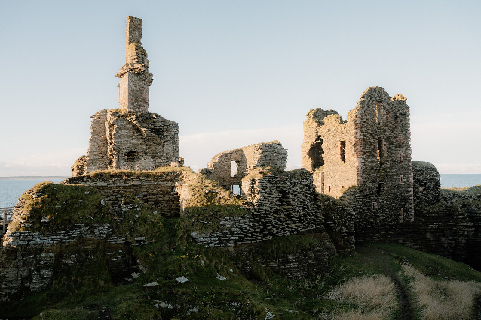 A ruined castle crowns a grassy headland, a broken chimney stack and roofless towers rising above the sea beyond.