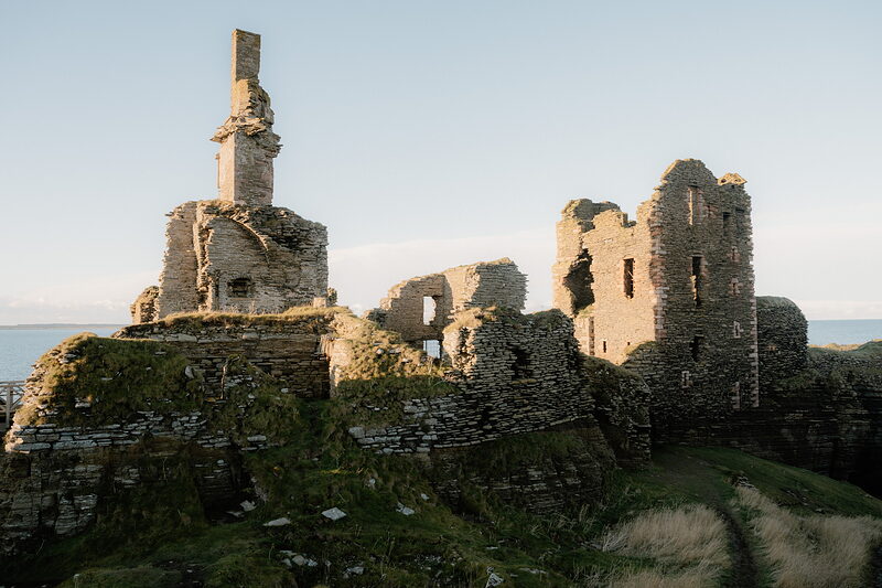 A ruined castle crowns a grassy headland, a broken chimney stack and roofless towers rising above the sea beyond.