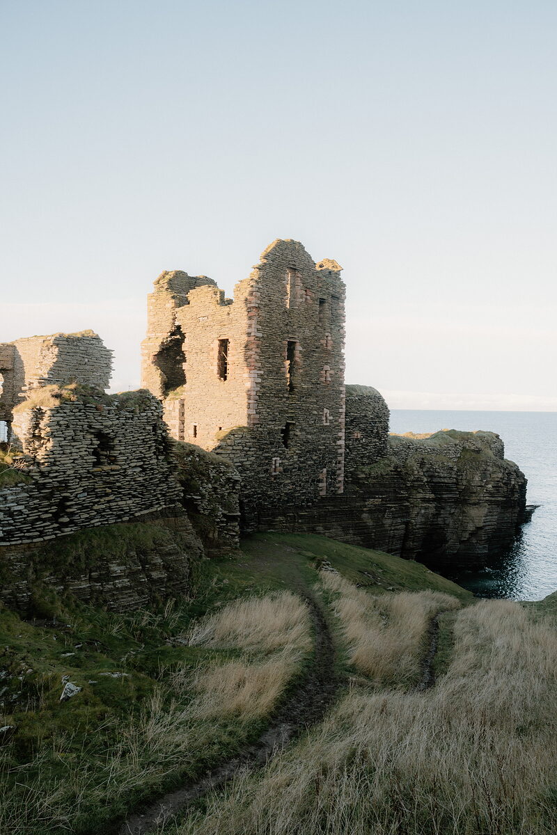 A ruined castle's tower and broken walls rise above a grassy headland, twin cart tracks leading towards the cliff edge and open sea beyond.