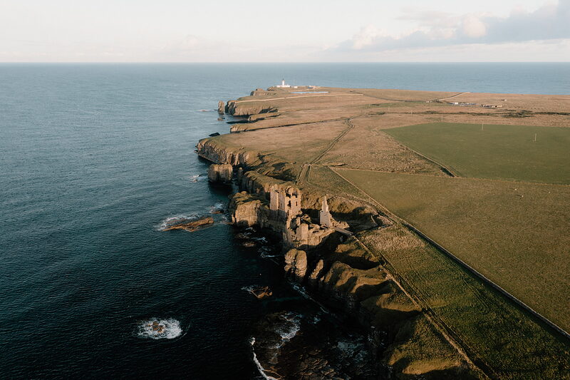 An aerial view shows a ruined castle perched on a narrow sea headland, cliffs dropping to the water and a lighthouse visible on the point beyond.