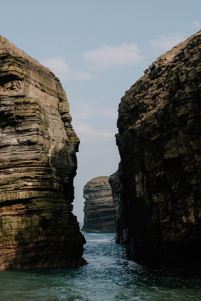 Two tall sea stacks of banded sedimentary rock frame a narrow gap of open sea, a smaller stack visible in the distance beyond them.