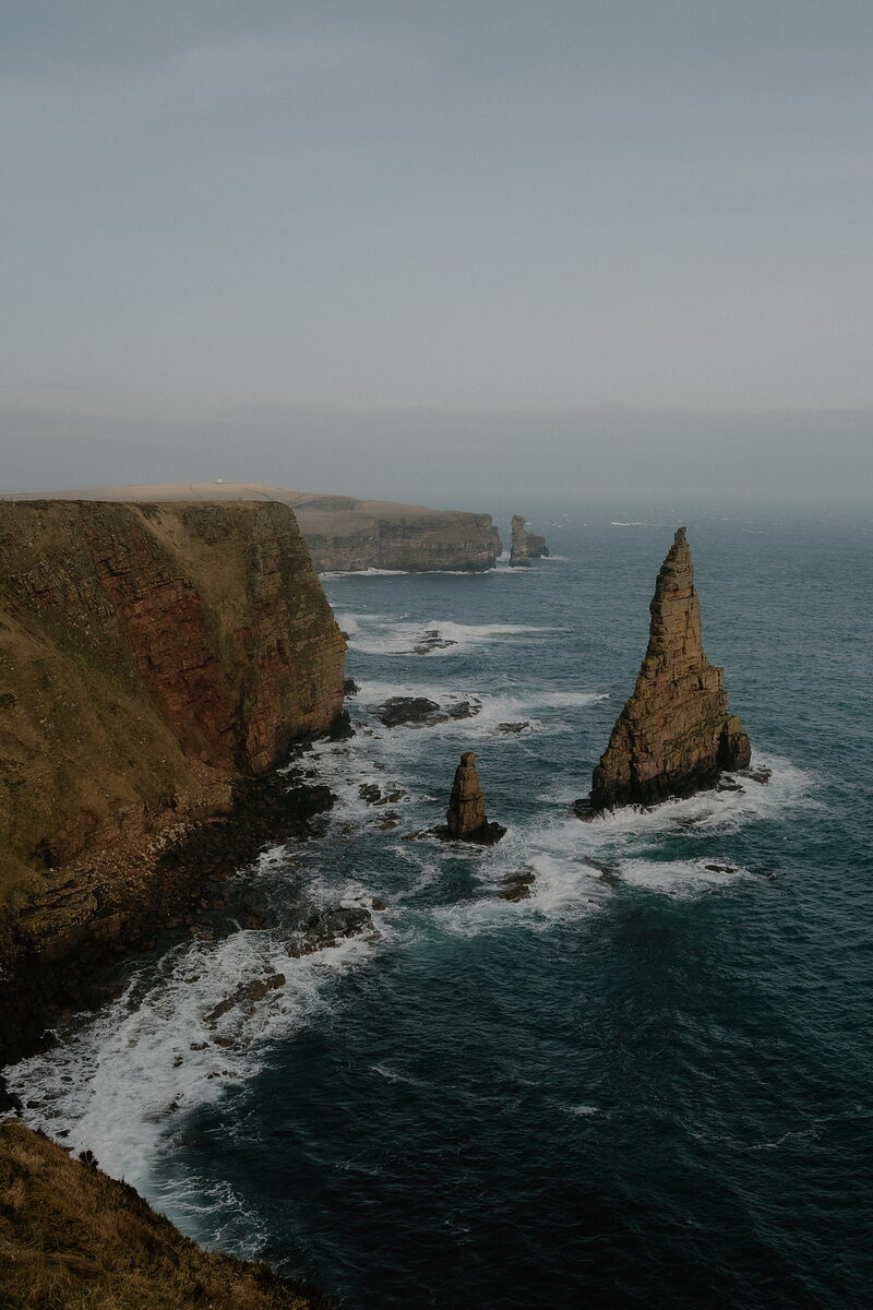A tall sea stack rises from the water beside layered cliffs, waves breaking white at its base under a hazy sky, a smaller stack visible behind.
