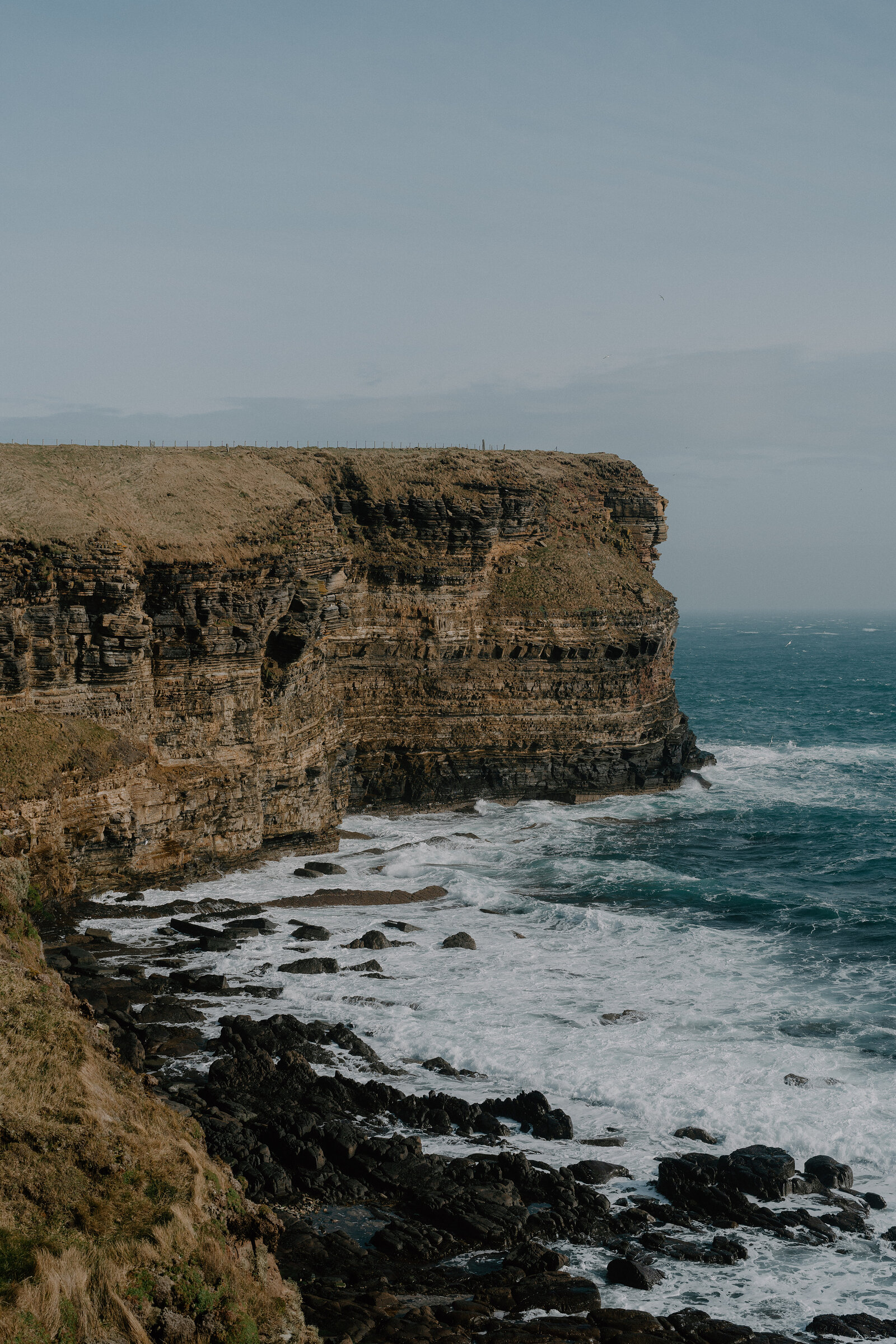 A high line of layered rock cliffs runs along the coast, waves breaking over rocks at the water's edge beneath a pale sky.