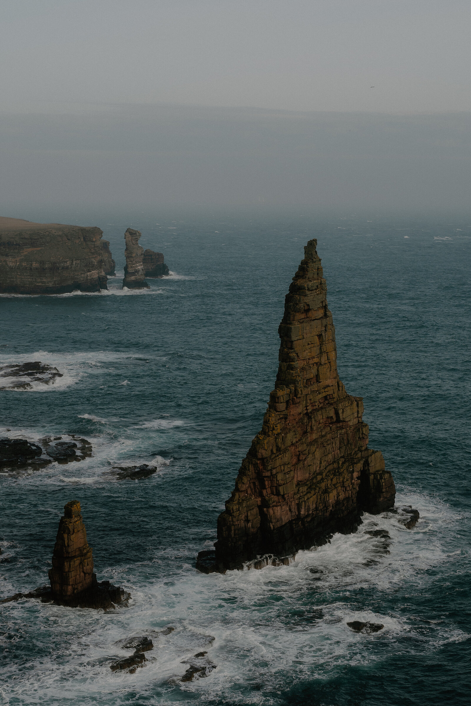 Two sea stacks rise from the water in the foreground, a further stack and headland visible in the haze beyond.