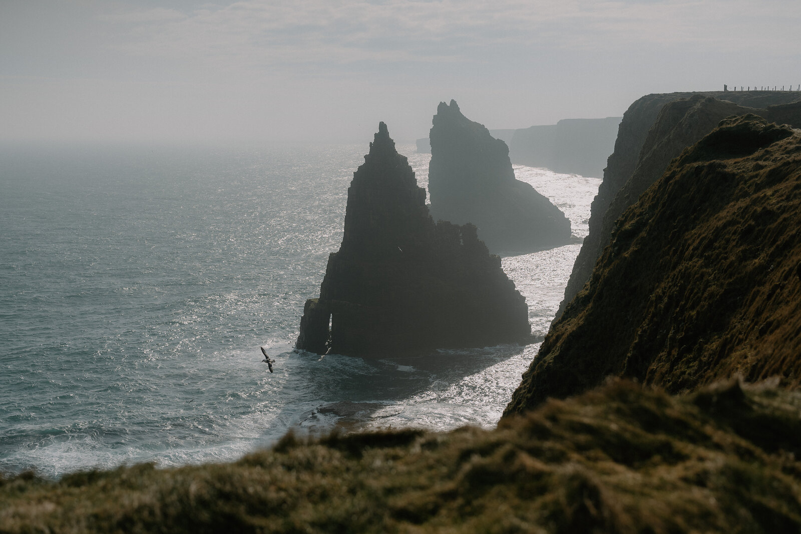 Two jagged sea stacks stand in silhouette below cliffs where onlookers stand at the edge, waves breaking around the stacks' bases in hazy light.