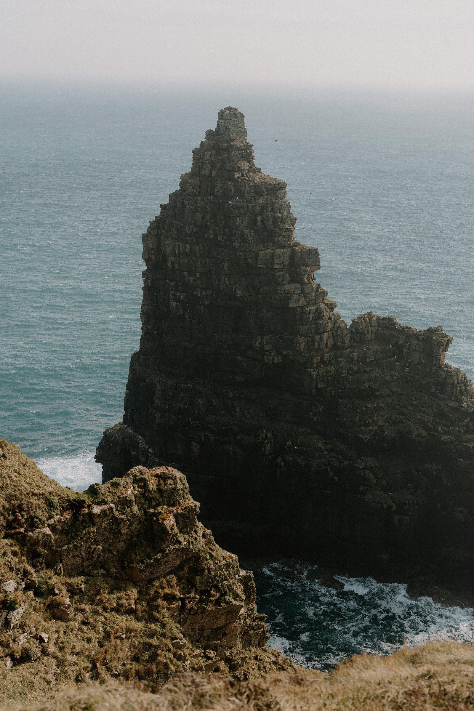A pointed sea stack rises steeply from the water beside a grassy cliff edge, waves breaking around its base in hazy light.