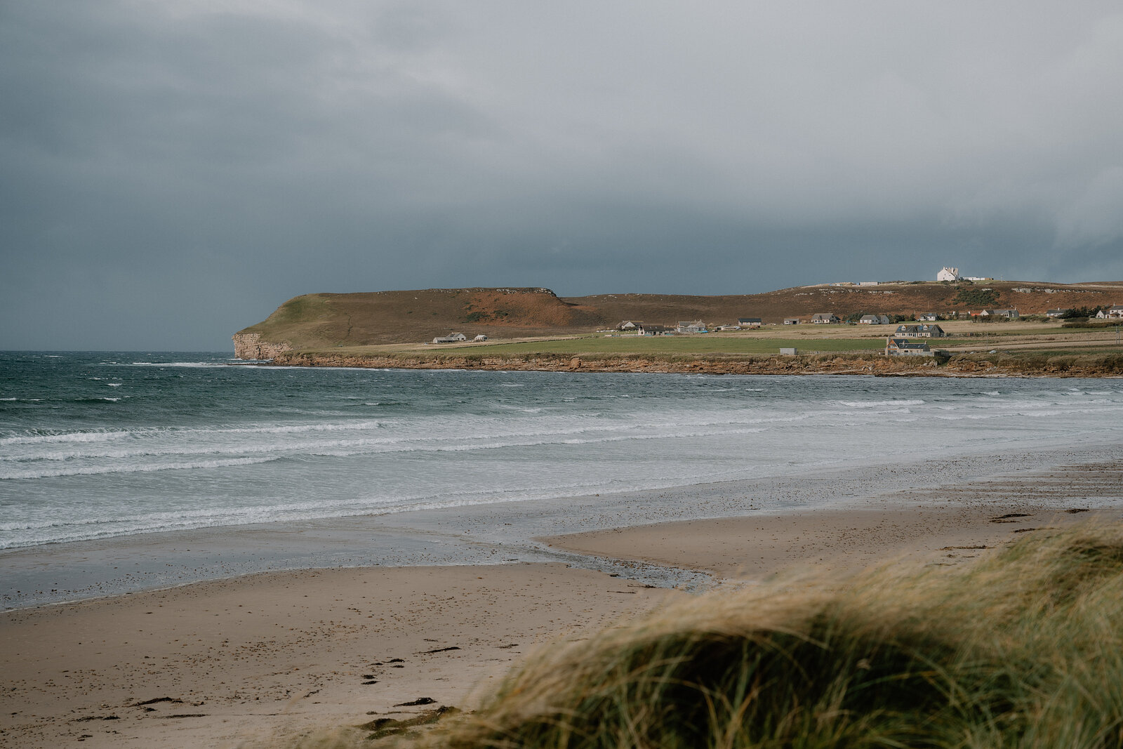 A wide sandy beach curves beneath a grassy headland dotted with houses, dune grass filling the foreground and waves rolling in under a stormy sky.