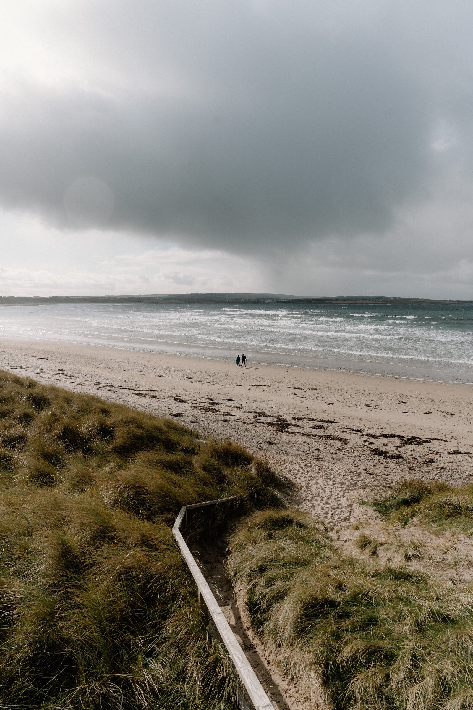 Two people walk along a wide sandy beach beneath a dramatic storm cloud, wooden steps descending through dune grass in the foreground.