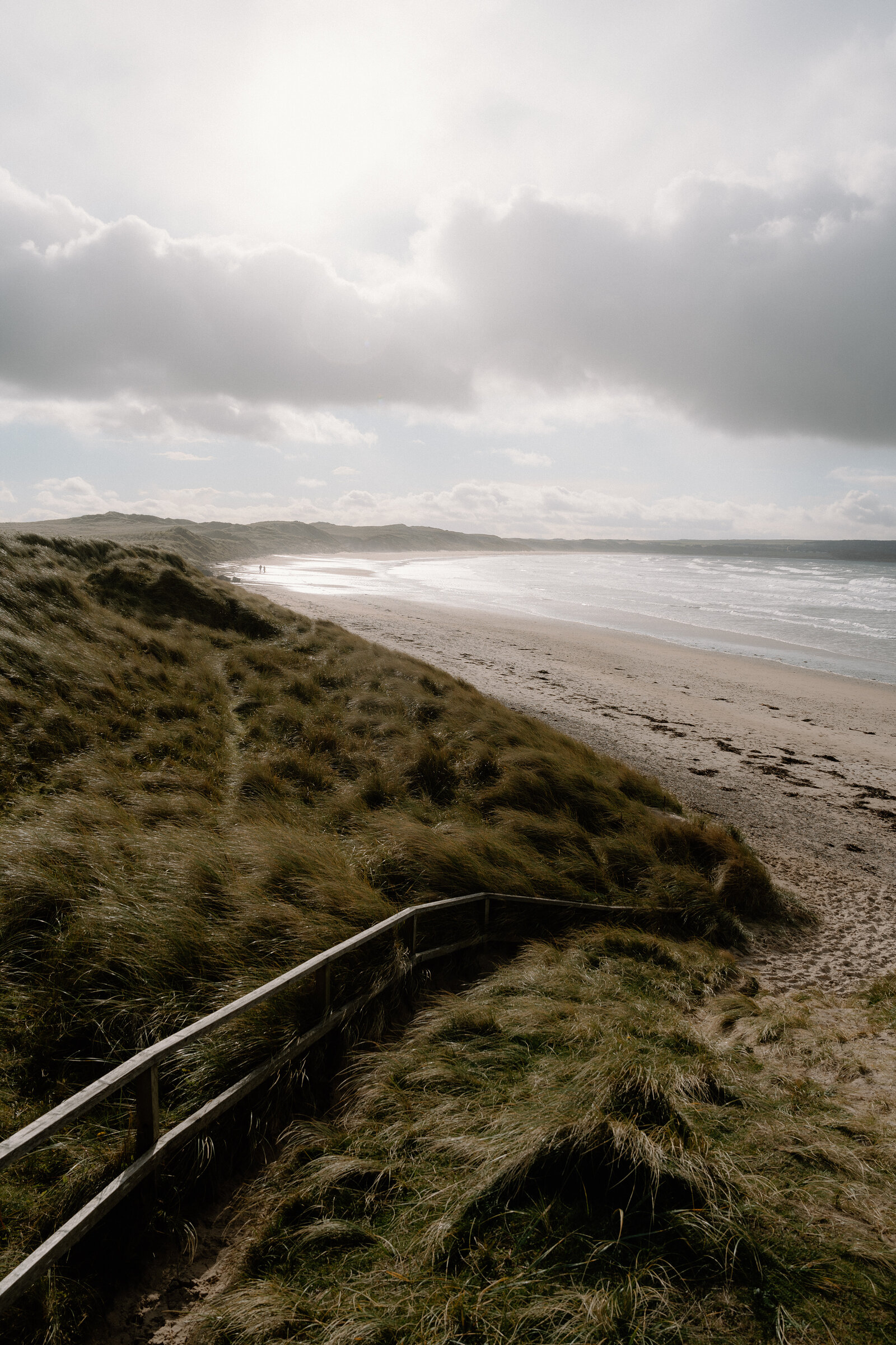A wooden railing follows a path through grassy dunes above a sandy beach where people walk near the water.