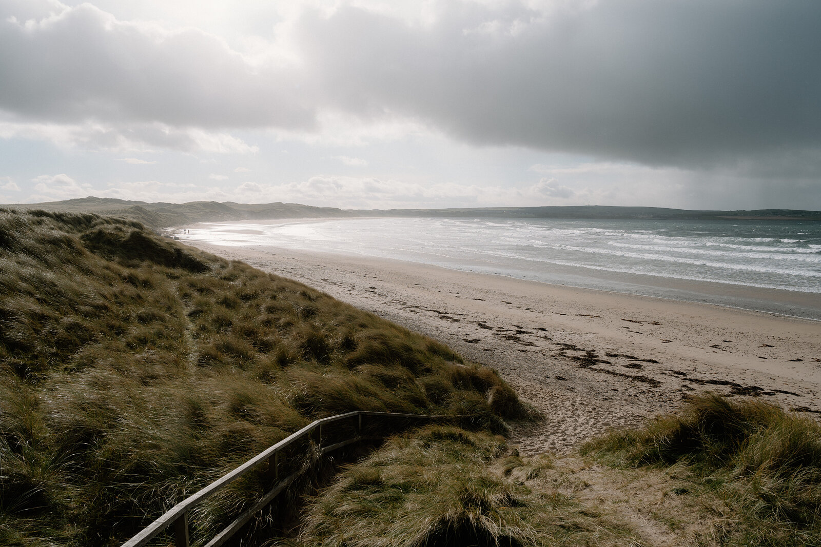 A wide sandy beach curves into the distance beneath dune grass and a cloudy sky, waves breaking gently along the shore.