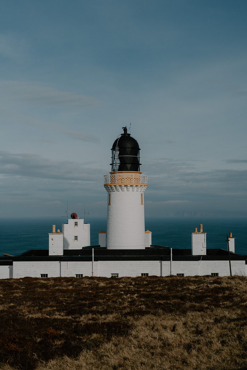 A white lighthouse with a black lantern room stands behind low outbuildings on a grassy clifftop, the sea stretching out under a dusky sky.
