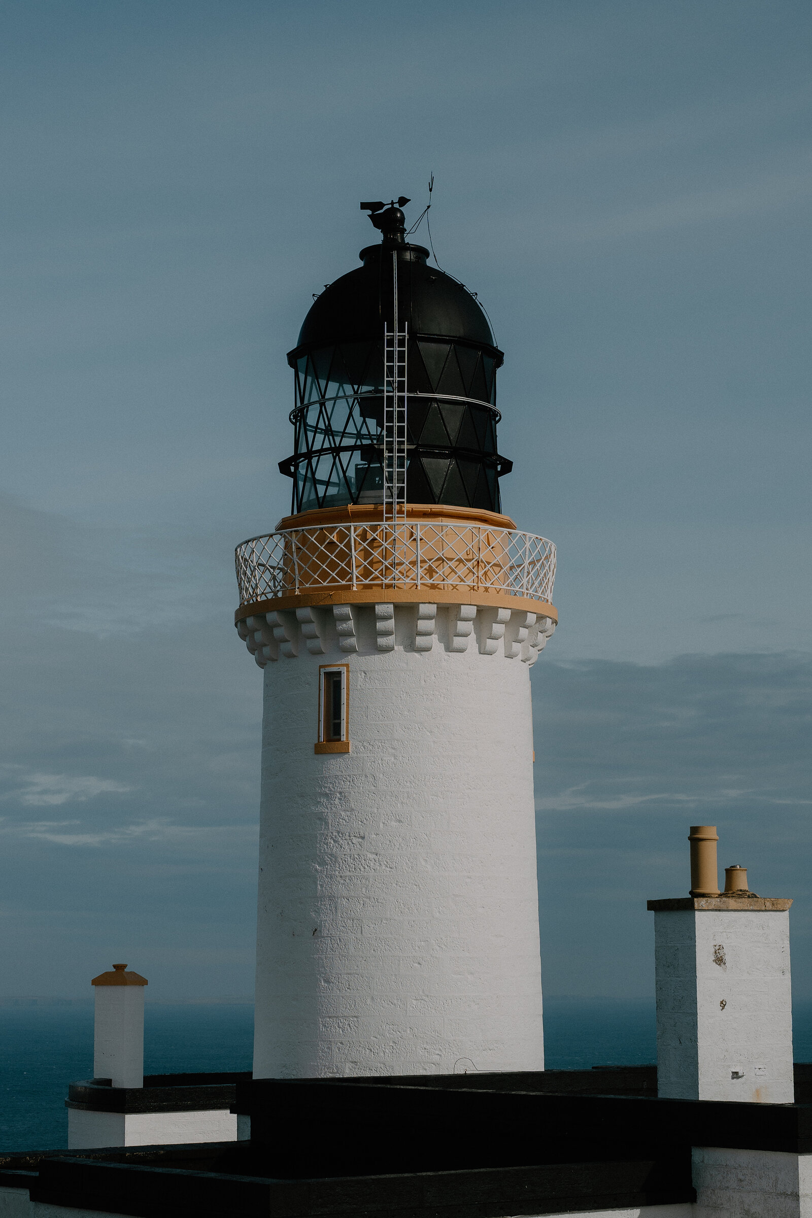 A close-up of a white lighthouse with a black lantern room and iron balcony, chimneys of low outbuildings visible below at dusk.