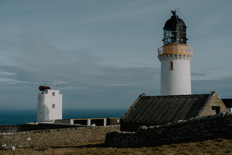 Two lighthouse buildings stand on a clifftop at dusk, a stone outbuilding with a slate roof in the foreground.