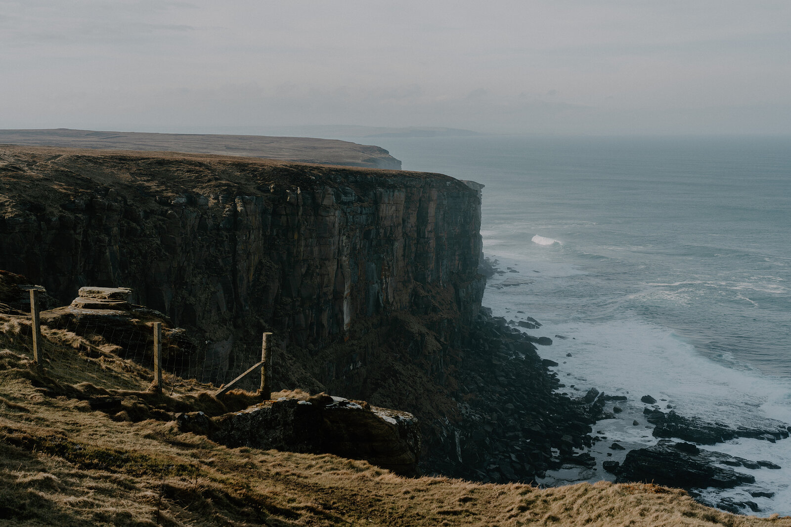 Tall dark cliffs stretch along a hazy coastline, wooden posts marking a path through the grass at the edge.