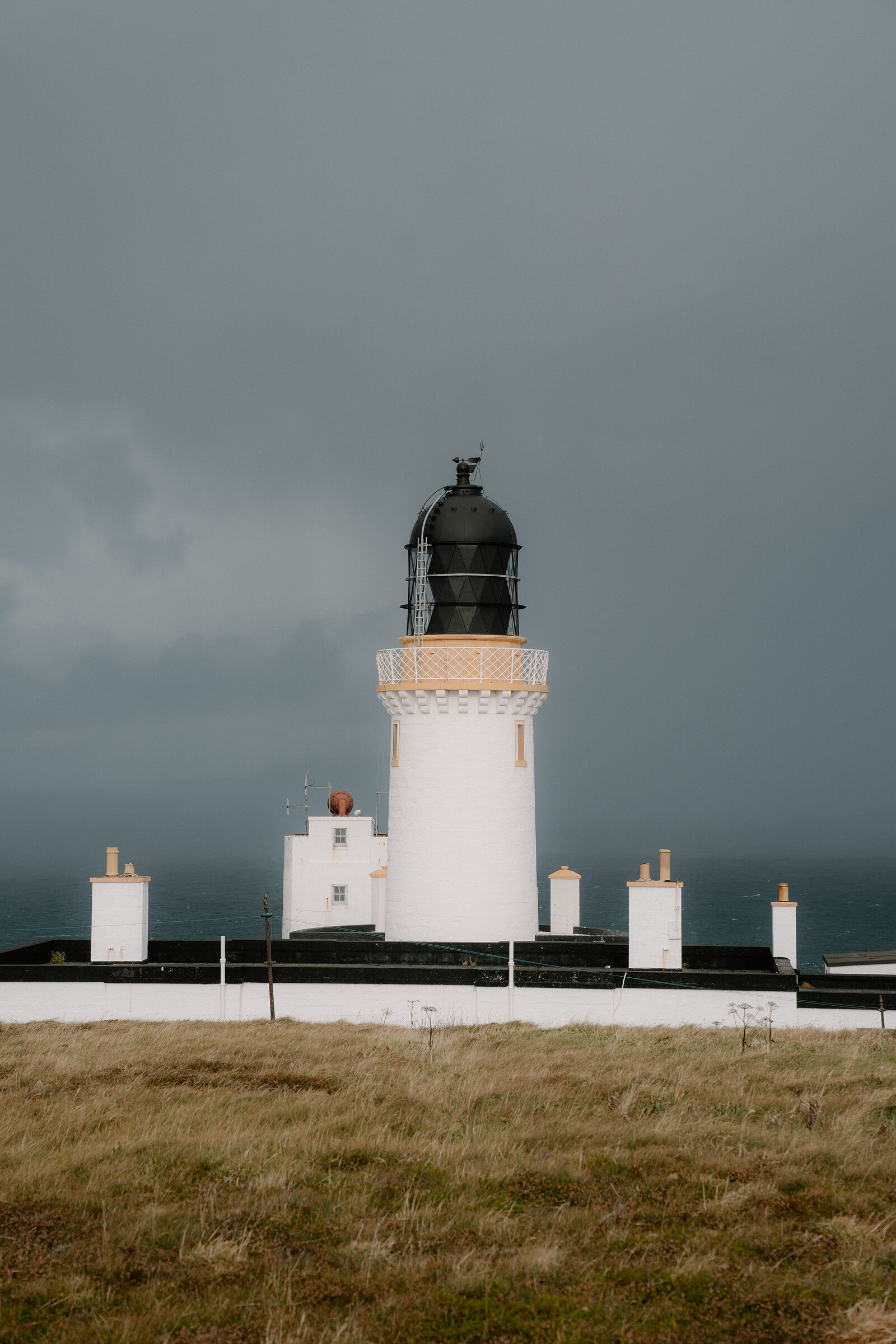 A white lighthouse with a black lantern room stands against a dark stormy sky, low outbuildings and grass in the foreground.