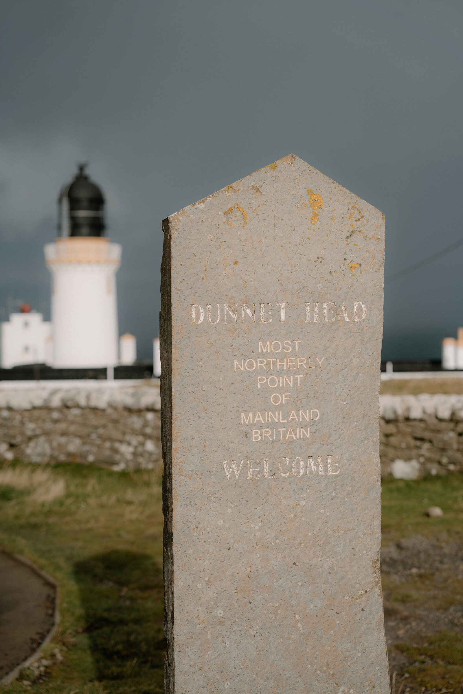 A stone marker reading "Dunnet Head, most northerly point of mainland Britain, welcome" stands before a lighthouse under a dark stormy sky.