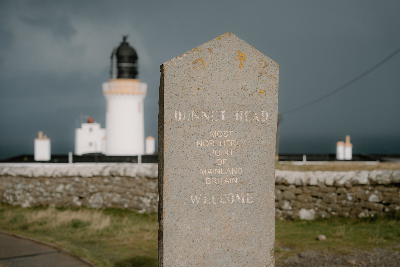 A stone marker reading "Dunnet Head, most northerly point of mainland Britain, welcome" stands before a lighthouse under a dark stormy sky.