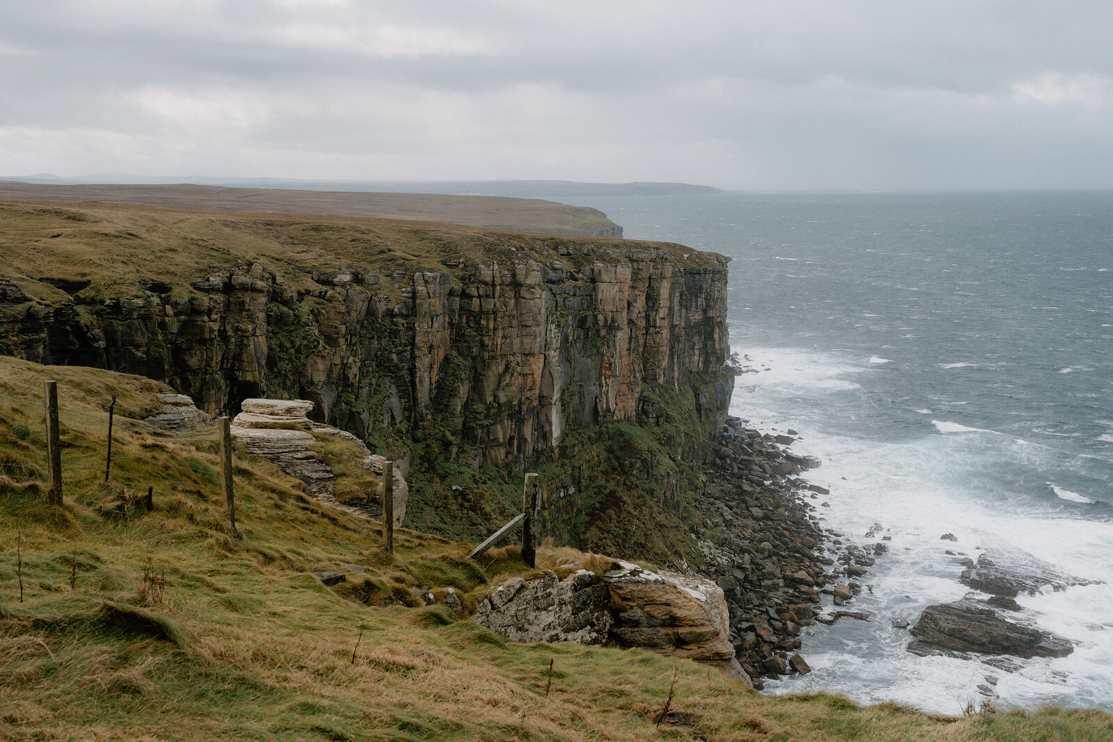 Tall dark cliffs stretch along a hazy coastline, waves breaking white at their base under a cloudy sky.