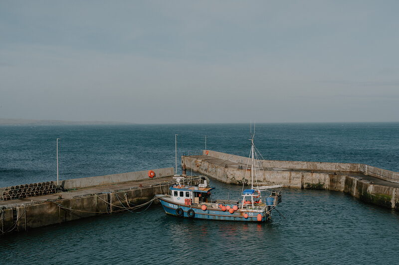 A blue fishing boat is moored inside a stone harbour, calm sea stretching beyond the breakwater walls.