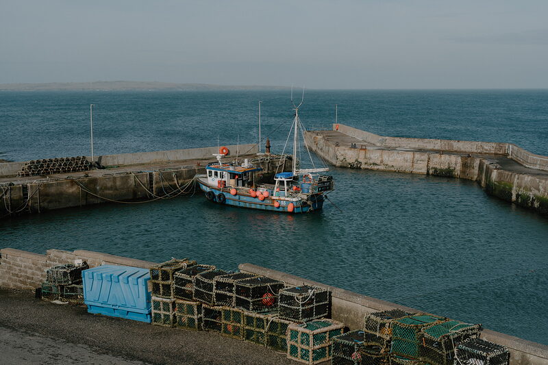 A blue fishing boat is moored inside a stone harbour, stacked lobster creels lining the quay in the foreground.