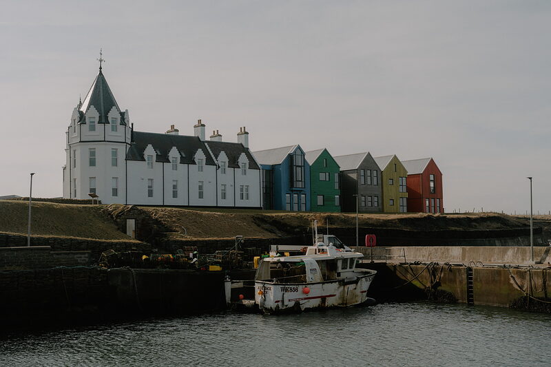 A turreted white castle-style hotel stands beside a row of colourful houses above a small harbour where a boat is moored.