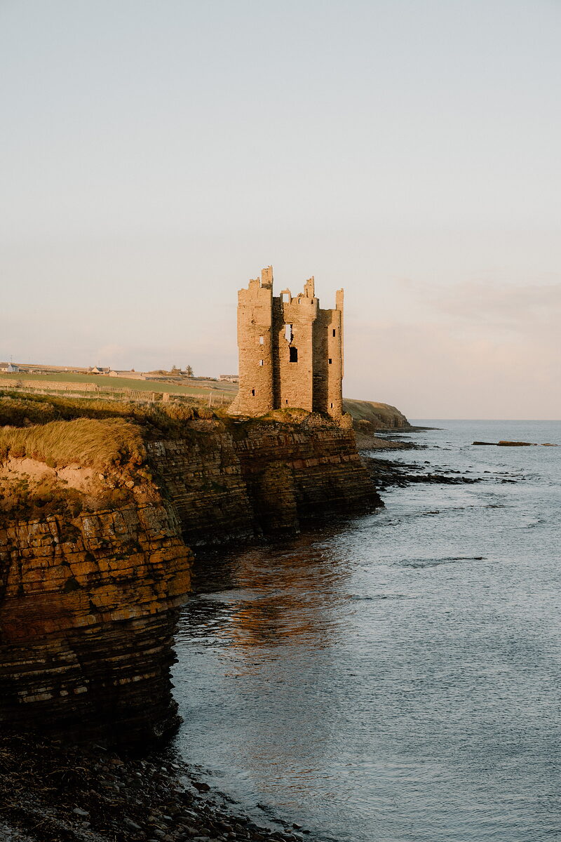 The ruined towers of a coastal castle stand on layered rock cliffs, warm evening light catching the stone above calm sea.