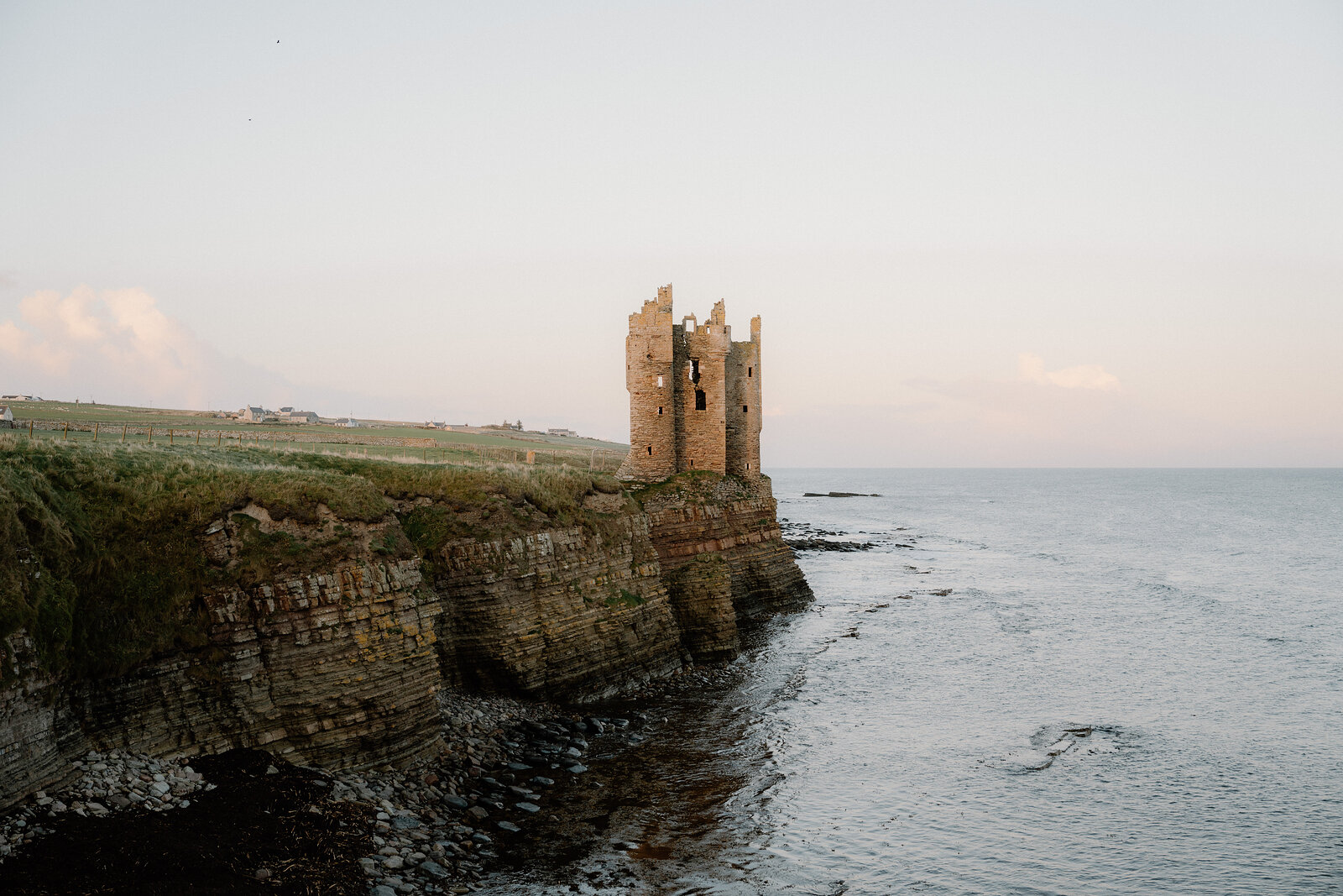 The roofless tower of Keiss Castle at the end of a layered rock cliff, a stony shore and calm sea below and fields running to the cliff top.