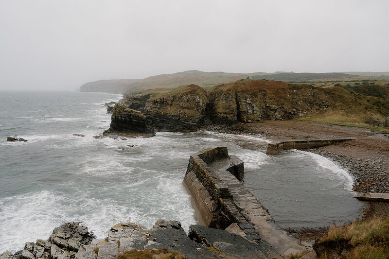 Waves surge into a walled harbour beneath layered sea cliffs, a stone pier curving out into the rough water.