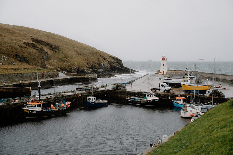 Small fishing boats crowd a stone harbour beneath a grassy headland, a white lighthouse standing at the harbour mouth.