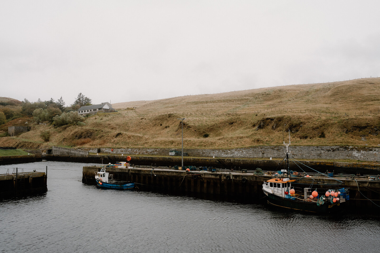 Two fishing boats are moored in a stone harbour beneath a grassy headland, a white cottage standing on the hillside above.