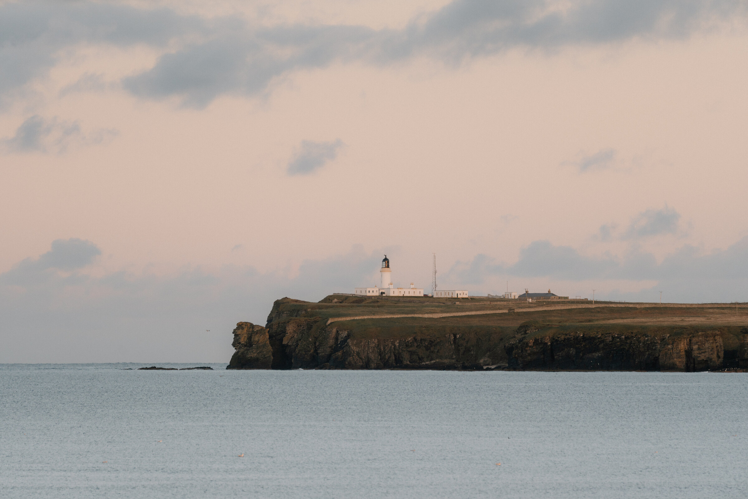 A distant lighthouse stands on a grassy headland above dark cliffs, calm sea stretching towards it under a soft evening sky.