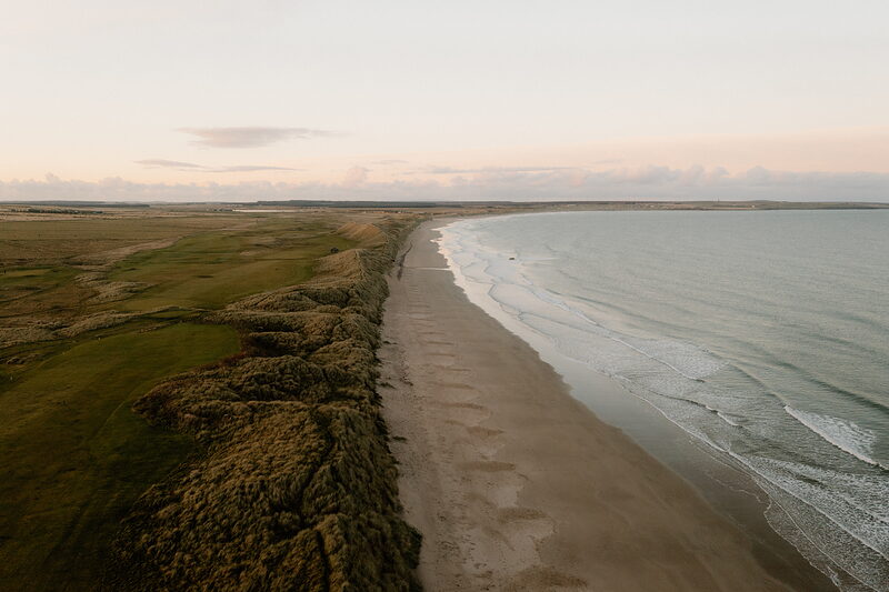 An aerial view follows a long sandy beach edged by dune grass at dusk, waves breaking gently along the shore.