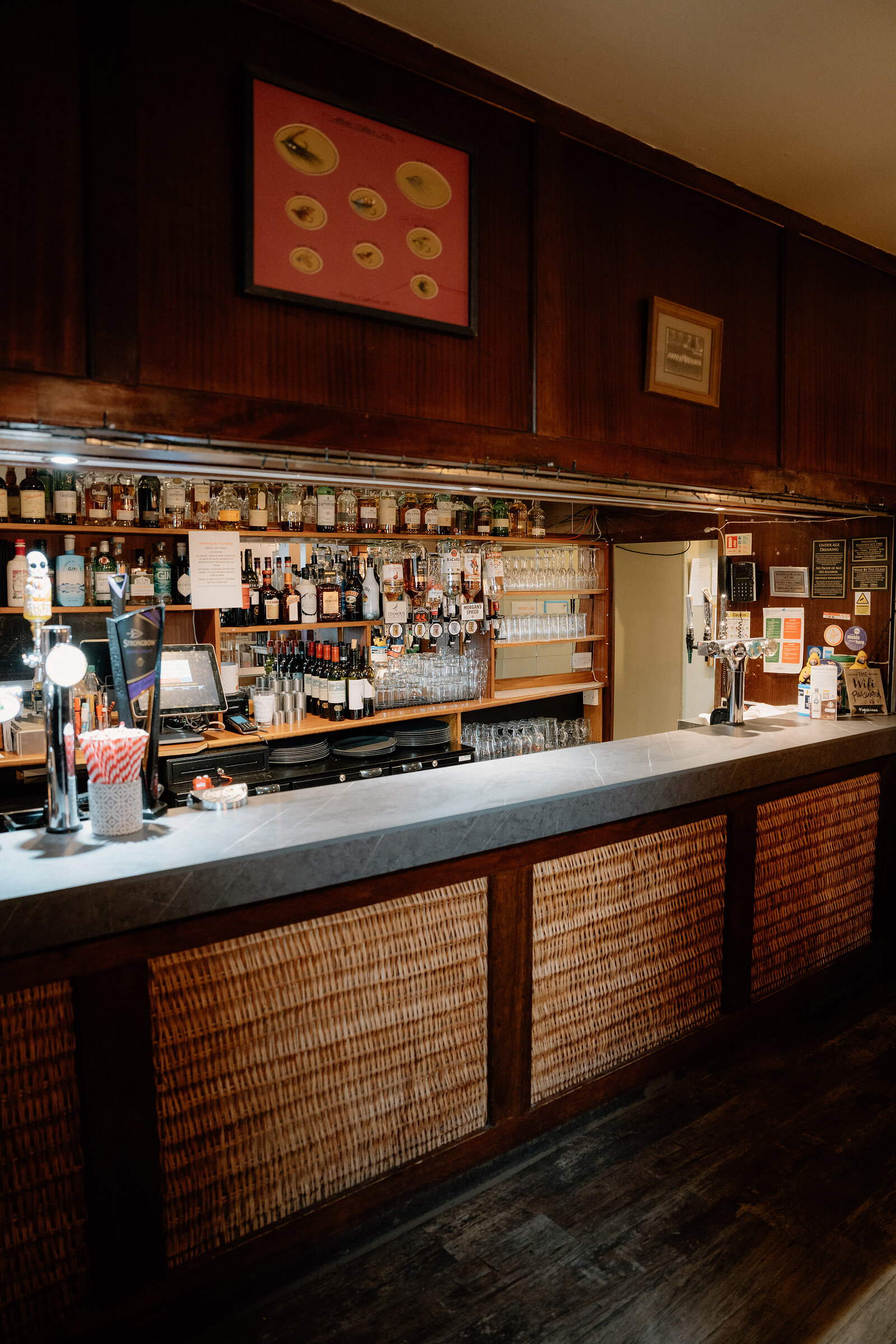 A dark wooden pub bar with wicker front panels stands beneath shelves of spirit bottles and beer taps, a framed print hanging above.