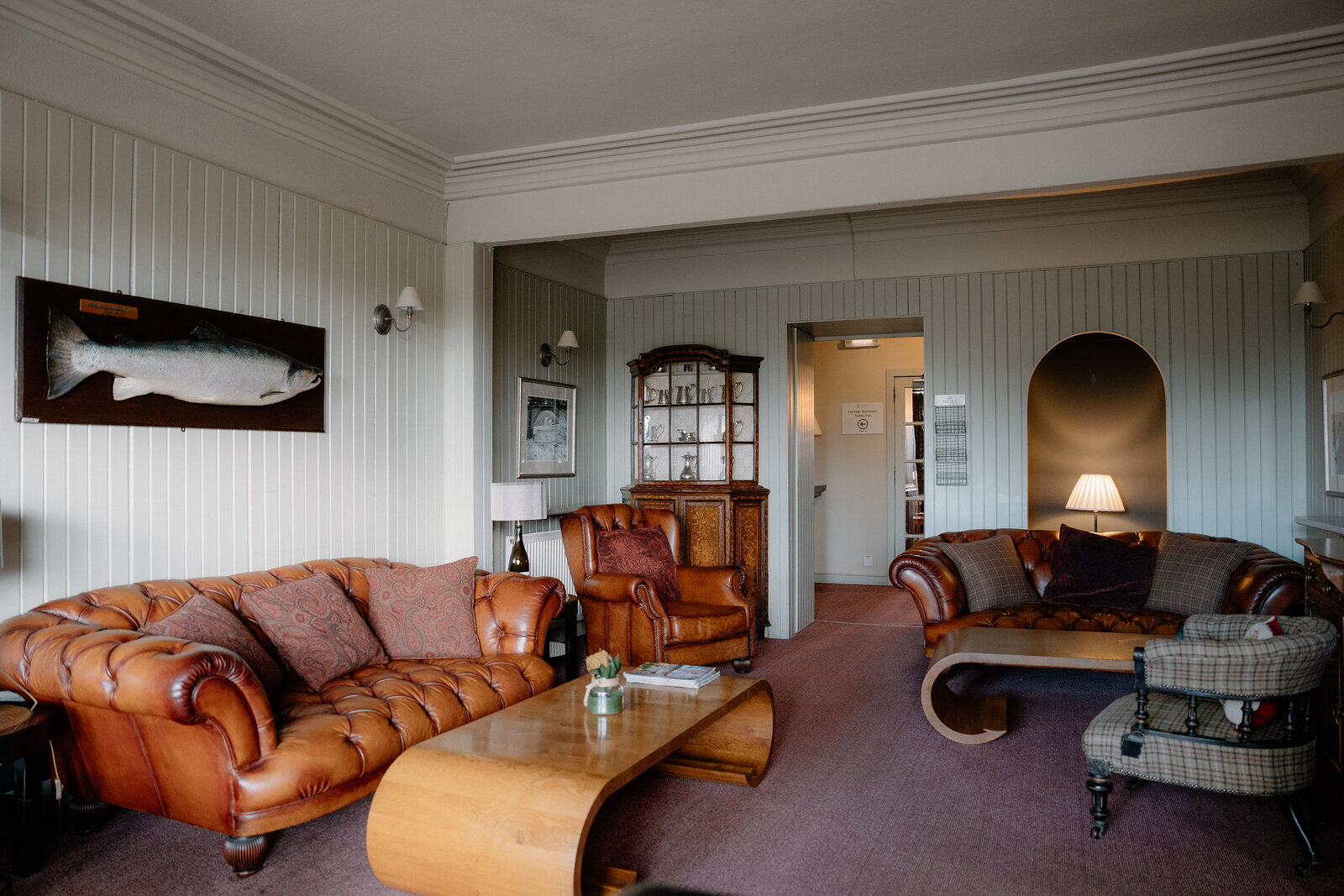 A pair of brown leather chesterfield sofas face each other across a low table in a panelled sitting room, a dark wood dresser and an arched alcove standing beyond, a framed fish print on the wall.