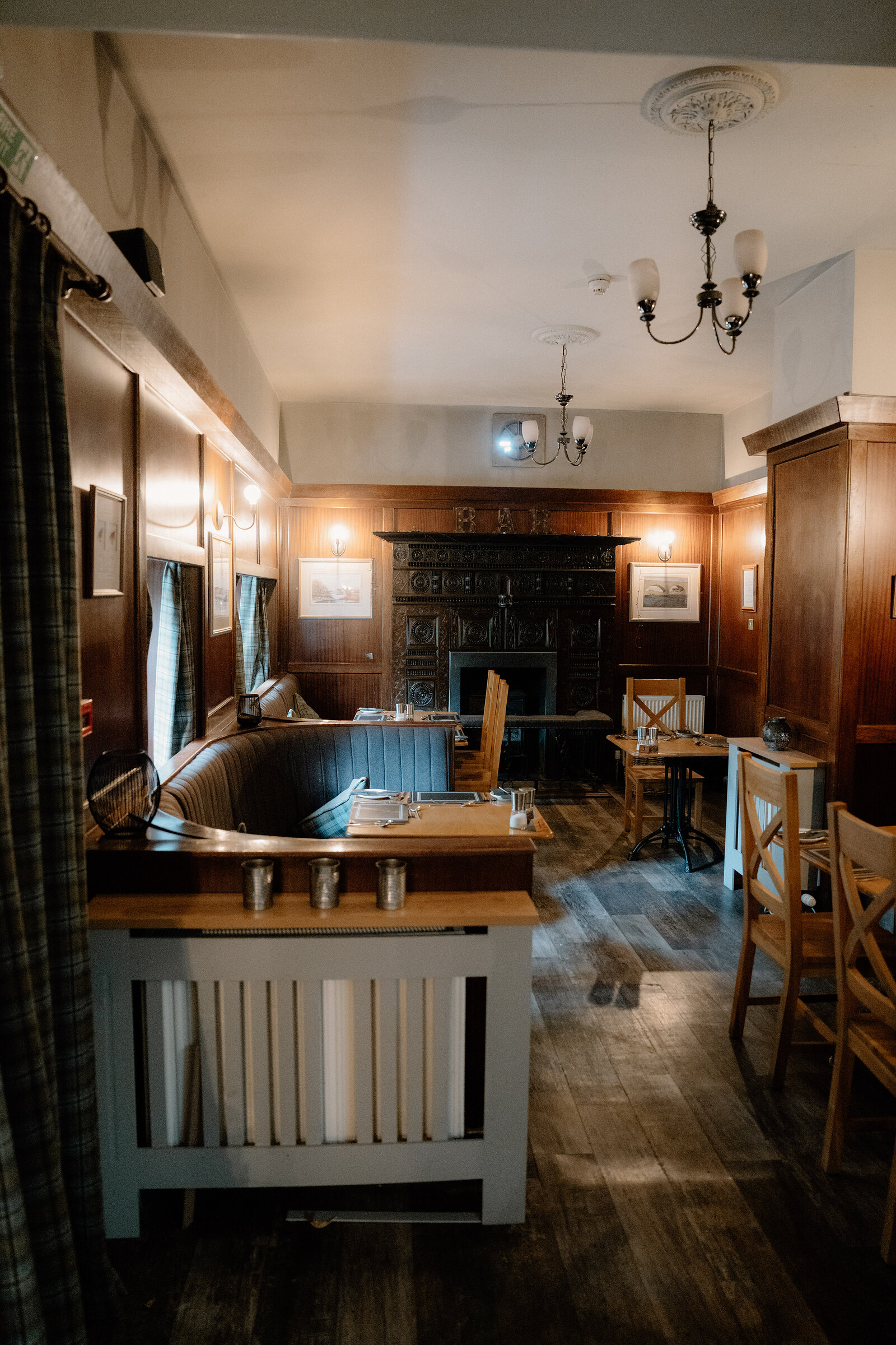 A wood-panelled dining nook with a moulded ceiling rose and chandelier holds a curved booth seat and small tables, a cast-iron fireplace visible beyond.