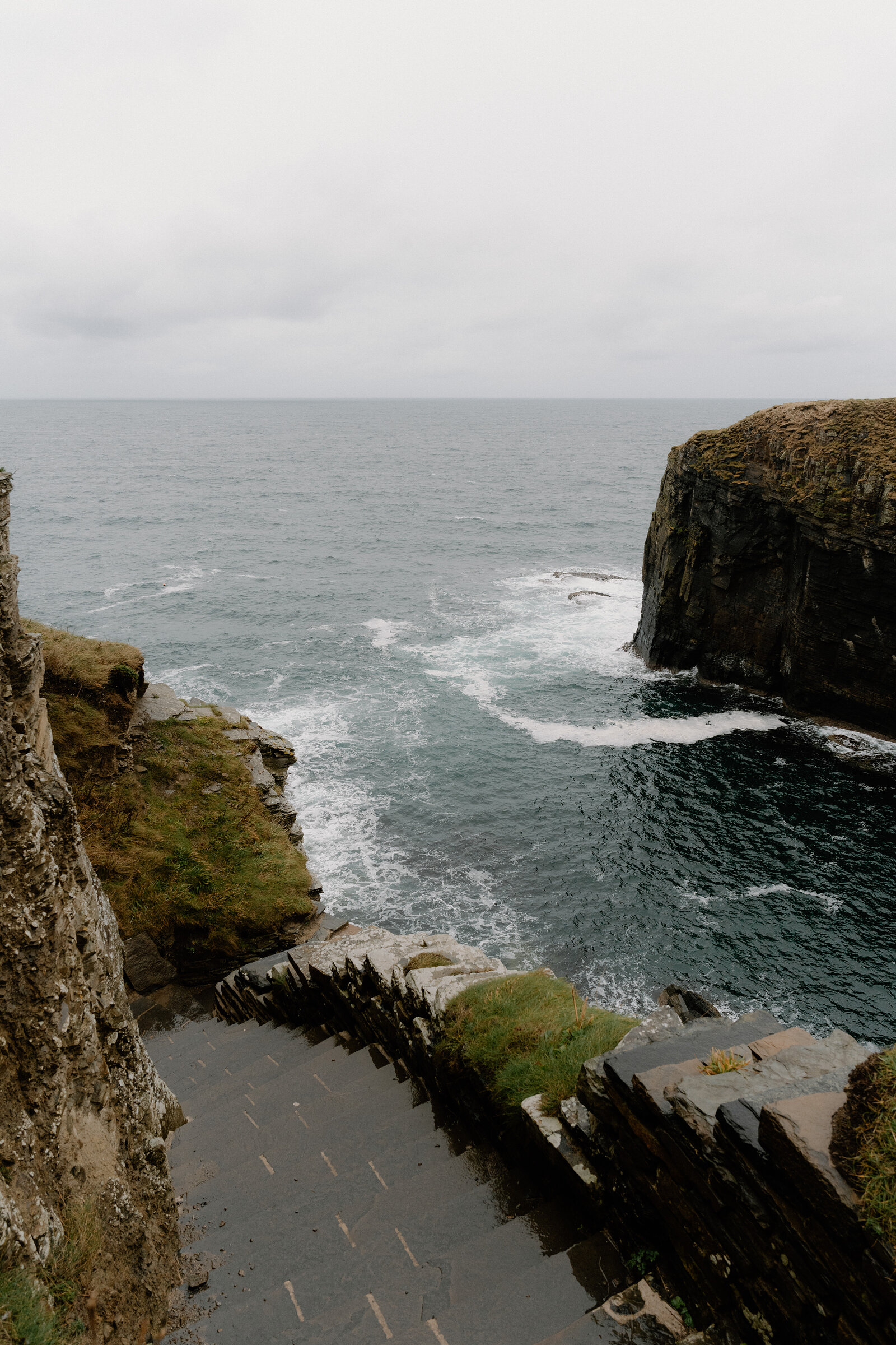 Stone steps descend to a churning sea inlet, waves breaking white against the cliffs on either side.