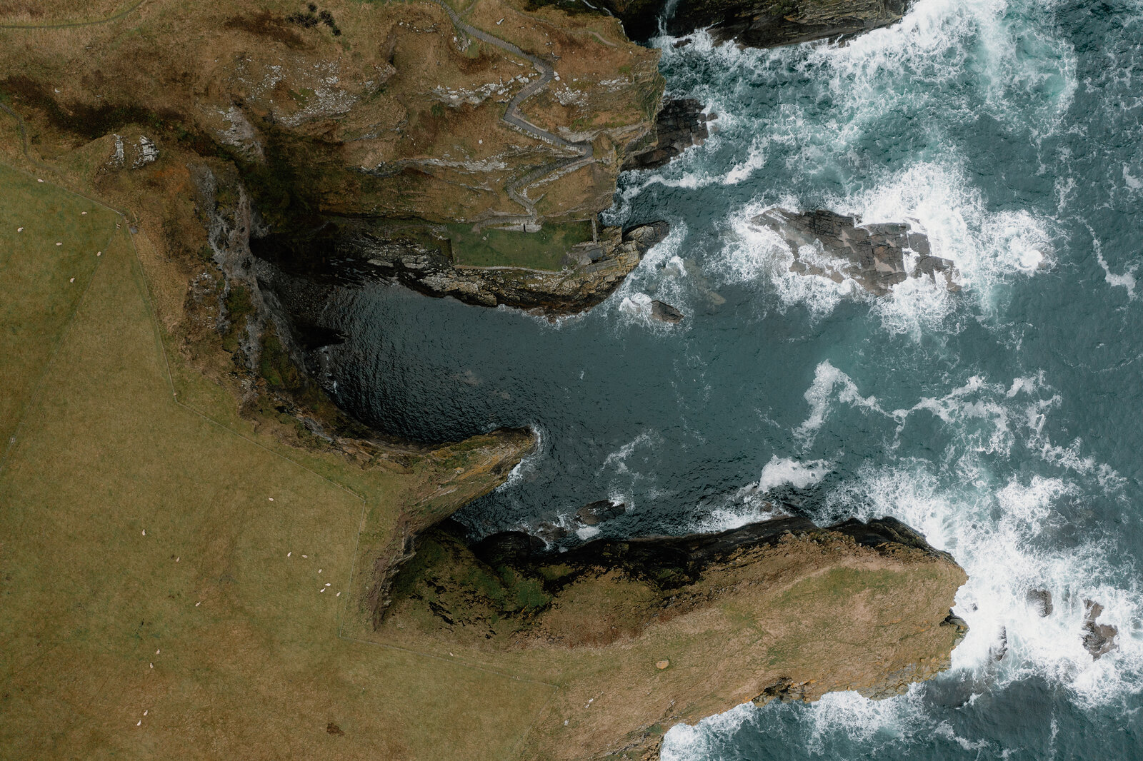 An aerial view shows a rocky coastline where a narrow sea inlet cuts into green clifftop fields, waves breaking white against the dark rocks.