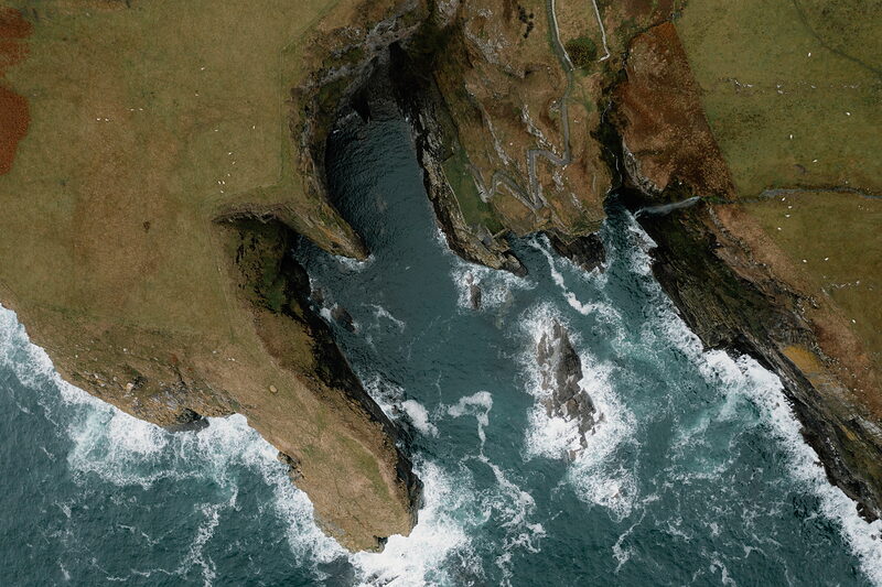 An aerial view shows a narrow sea channel winding between rocky headlands, waves breaking white where the channel meets open water, and a path running along the clifftop above.