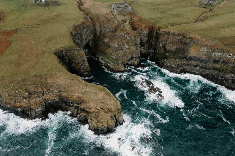 An aerial view shows a rugged headland where dark cliffs meet a turquoise sea, waves breaking white against sea stacks and a narrow inlet cutting into the rock.