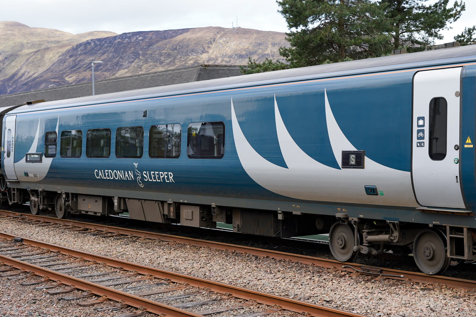 A Caledonian Sleeper carriage in its dark blue livery stands on the track with Fort William on its destination board, heather hills rising behind.