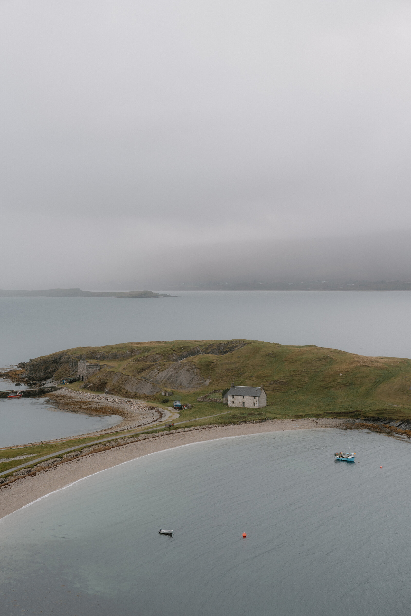 A grassy headland reaches into a calm bay, a white cottage standing on the point and small boats anchored in the misty water.