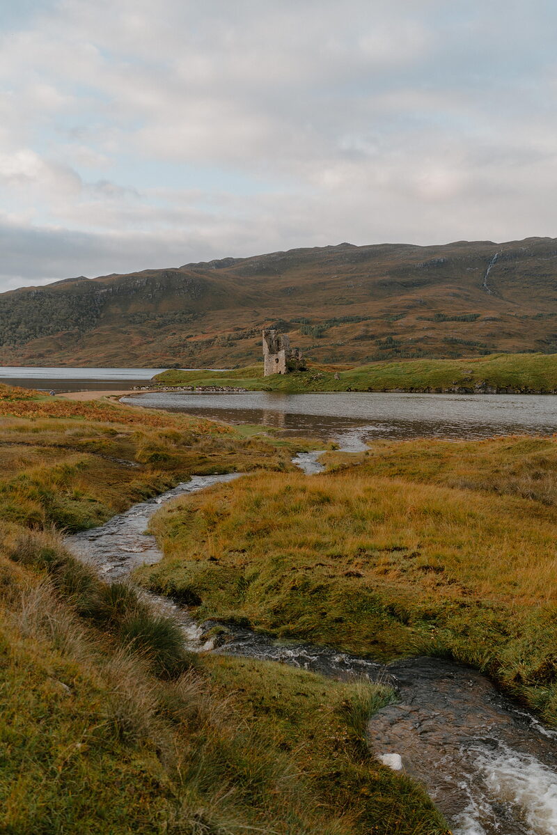 A ruined castle stands on a small headland surrounded by a loch, a stream winding through golden autumn grass towards the water.