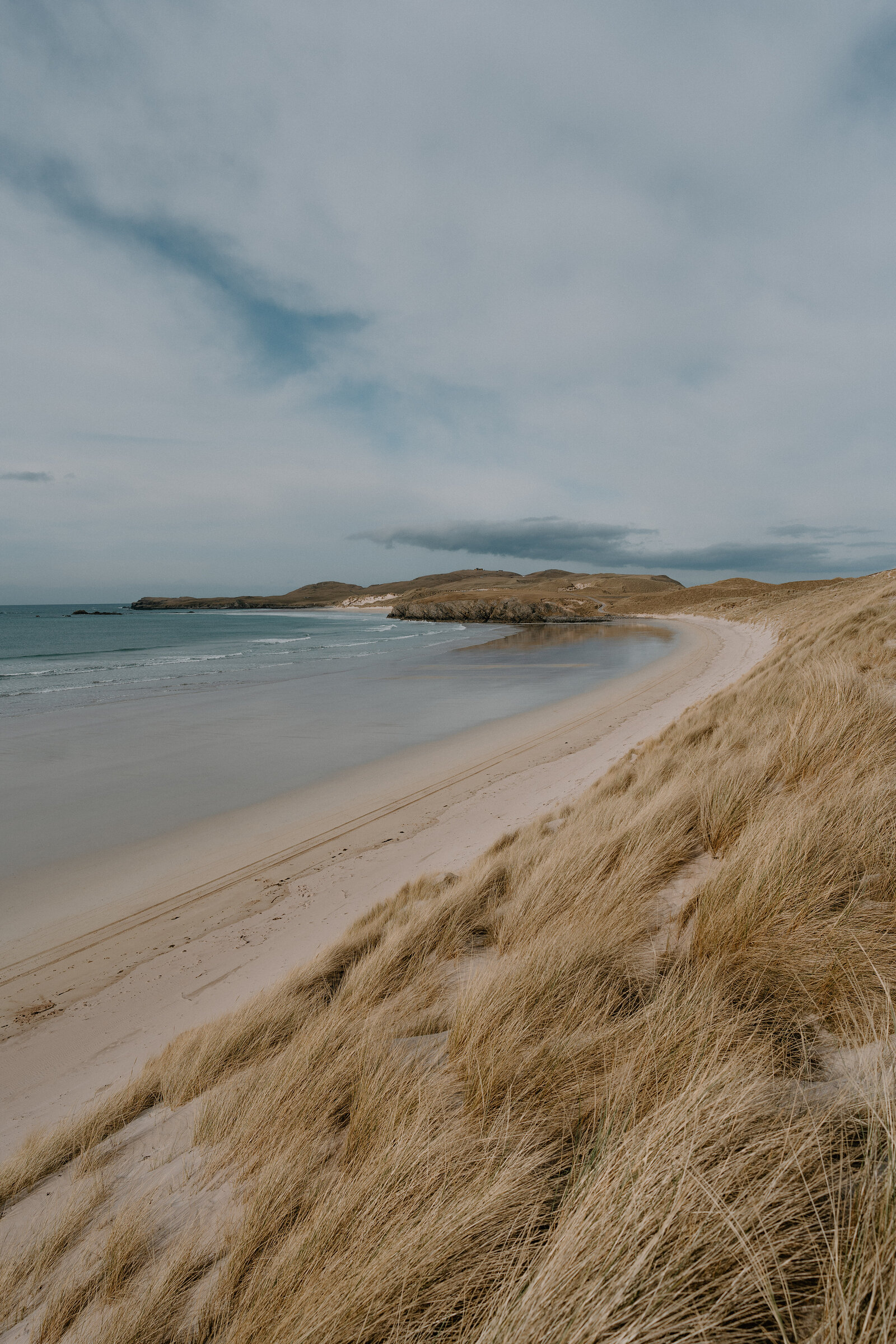 A curving sandy beach meets calm sea below a rocky headland, marram grass covering the dunes in the foreground.
