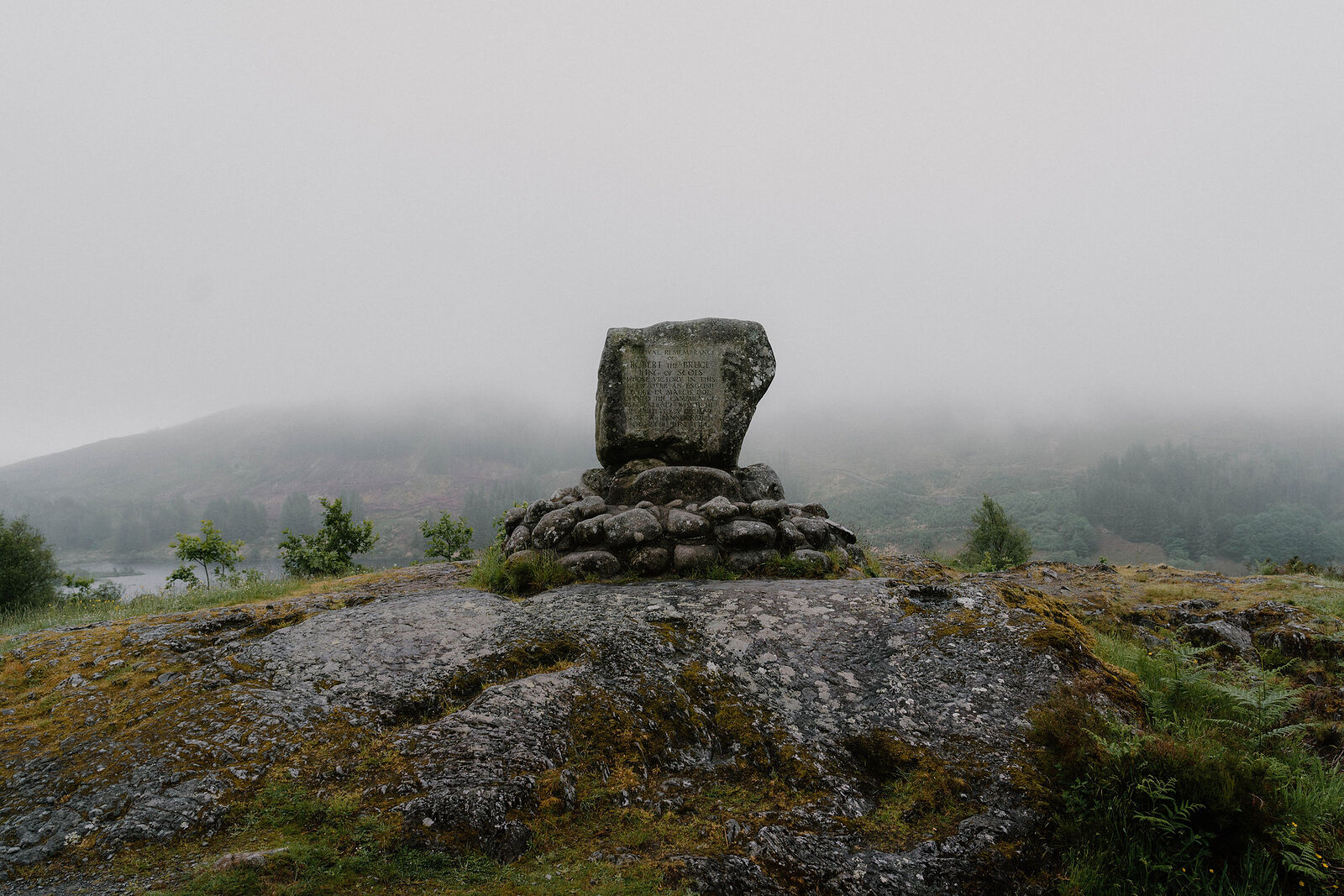 A stone cairn topped with a flat slab stands on a mossy rock outcrop, mist obscuring the hills behind.