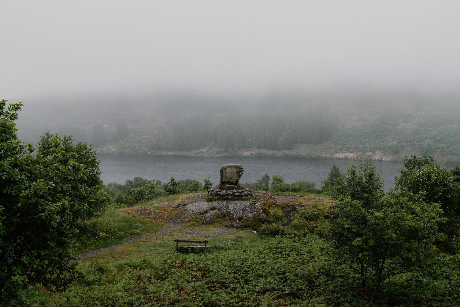 A stone monument stands on a hillside above a misty loch, a bench and gravel path nearby and trees framing the view.